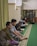 Several people are sitting on a green carpeted floor in a room that appears to be a mosque or a prayer room. Some are reading from books, while others sit quietly. The room has a bookshelves filled with books and a curtain on one side.