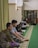 Several people are sitting on a green carpeted floor in a room that appears to be a mosque or a prayer room. Some are reading from books, while others sit quietly. The room has a bookshelves filled with books and a curtain on one side.