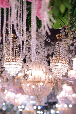 Opulent crystal chandeliers with intricate gold detailing hang elegantly among lush greenery and floral decorations. The scene is illuminated with soft, warm lighting that creates a dreamy and luxurious atmosphere.