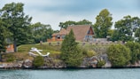 A picturesque lakefront property featuring an A-frame house surrounded by lush greenery. The house is situated on a grassy hill with several trees and a prominent white anchor decoration on the slope. The scene is calm and serene, with a body of water in the foreground and a cloudy sky above.