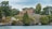 A picturesque lakefront property featuring an A-frame house surrounded by lush greenery. The house is situated on a grassy hill with several trees and a prominent white anchor decoration on the slope. The scene is calm and serene, with a body of water in the foreground and a cloudy sky above.