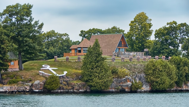 A picturesque lakefront property featuring an A-frame house surrounded by lush greenery. The house is situated on a grassy hill with several trees and a prominent white anchor decoration on the slope. The scene is calm and serene, with a body of water in the foreground and a cloudy sky above.