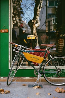A vintage-style bicycle with a yellow banner advertising Jameson Irish Whiskey is parked outside a storefront. The shop window displays a warm interior with decorative elements, and an 'Open' sign hangs on the green exterior wall. Fallen leaves are scattered on the ground, and trees with bare branches are reflected in the glass.