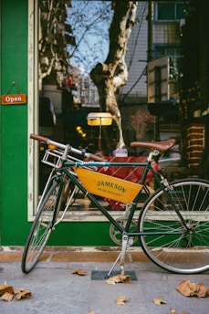 A vintage-style bicycle with a yellow banner advertising Jameson Irish Whiskey is parked outside a storefront. The shop window displays a warm interior with decorative elements, and an 'Open' sign hangs on the green exterior wall. Fallen leaves are scattered on the ground, and trees with bare branches are reflected in the glass.