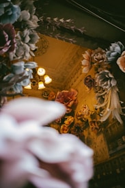 An interior space is lavishly decorated with large, artificial flowers in soft pastel colors like pink, white, and peach. The ceiling features ornate molding and a vintage chandelier with warm lighting. The walls are adorned with more flowers, adding a dreamy and whimsical atmosphere.