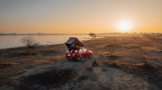A family enjoying breakfast beside their car with a rooftop tent set up in a sunny meadow.