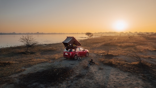 A red car with a rooftop tent is parked on a flat, grassy landscape by a lake. The sun is setting or rising, casting a warm golden glow over the area. In the distance, there are some scattered trees and a hazy horizon.