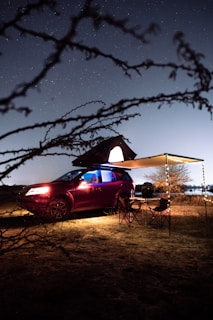 A red SUV is parked on a sandy ground at night, with a rooftop tent deployed. An awning extends from the side, sheltering a small camp setup with foldable chairs and a table. The scene is dimly lit, with strings of lights providing ambient illumination. Starry night sky and silhouetted tree branches frame the image, adding to the tranquil outdoor setting.