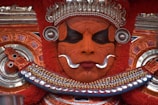 A person adorned in traditional Theyyam attire, featuring intricate facial makeup with vibrant orange and black patterns. The look is complemented by elaborate silver and red headdress and jewelry, with detailed carvings and embellishments enhancing the mystical and regal appearance.