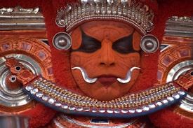 A person adorned in traditional Theyyam attire, featuring intricate facial makeup with vibrant orange and black patterns. The look is complemented by elaborate silver and red headdress and jewelry, with detailed carvings and embellishments enhancing the mystical and regal appearance.