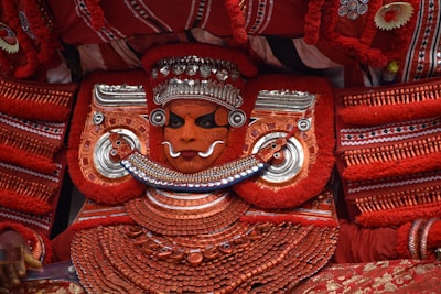 An elaborate traditional costume features a person adorned in intricate red and silver attire, with detailed face paint and ornate jewelry. The costume includes large decorative elements and patterns, creating a striking and vibrant appearance.