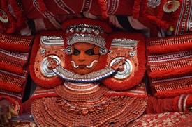 An elaborate traditional costume features a person adorned in intricate red and silver attire, with detailed face paint and ornate jewelry. The costume includes large decorative elements and patterns, creating a striking and vibrant appearance.