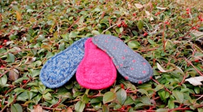 Three fabric pads with different patterns and colors, including blue with floral prints, pink, and grey with dots, are placed on lush green foliage with small red berries.