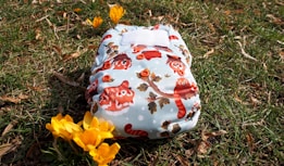A cloth diaper with a colorful pattern featuring cartoon animals, resting on grassy ground with fallen leaves around. Bright yellow flowers are nearby, adding a touch of spring to the scene.