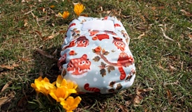 A cloth diaper with a colorful pattern featuring cartoon animals, resting on grassy ground with fallen leaves around. Bright yellow flowers are nearby, adding a touch of spring to the scene.