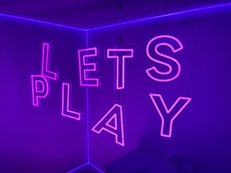 Neon letters spelling 'LETS PLAY' are suspended in a room bathed in purple light, creating a vibrant and playful atmosphere.