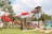 A vibrant outdoor playground featuring multiple slides, including a prominent red tube slide structure. The area is surrounded by lush greenery, including a tree and grass. A pool is visible near the base of one of the slides.