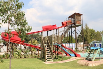 A vibrant outdoor playground featuring multiple slides, including a prominent red tube slide structure. The area is surrounded by lush greenery, including a tree and grass. A pool is visible near the base of one of the slides.