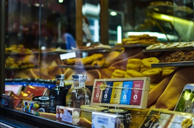 A shop window displays a variety of gourmet food items, including bottles of alcohol, neatly packaged chocolates, and tins. Golden pastries and cakes are arranged prominently on tiered shelves inside the window. The background features warm, inviting lighting that highlights the delicacies.