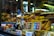 A shop window displays a variety of gourmet food items, including bottles of alcohol, neatly packaged chocolates, and tins. Golden pastries and cakes are arranged prominently on tiered shelves inside the window. The background features warm, inviting lighting that highlights the delicacies.