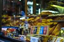 A shop window displays a variety of gourmet food items, including bottles of alcohol, neatly packaged chocolates, and tins. Golden pastries and cakes are arranged prominently on tiered shelves inside the window. The background features warm, inviting lighting that highlights the delicacies.