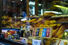A shop window displays a variety of gourmet food items, including bottles of alcohol, neatly packaged chocolates, and tins. Golden pastries and cakes are arranged prominently on tiered shelves inside the window. The background features warm, inviting lighting that highlights the delicacies.