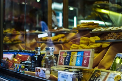 A shop window displays a variety of gourmet food items, including bottles of alcohol, neatly packaged chocolates, and tins. Golden pastries and cakes are arranged prominently on tiered shelves inside the window. The background features warm, inviting lighting that highlights the delicacies.