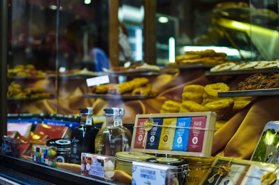 A shop window displays a variety of gourmet food items, including bottles of alcohol, neatly packaged chocolates, and tins. Golden pastries and cakes are arranged prominently on tiered shelves inside the window. The background features warm, inviting lighting that highlights the delicacies.