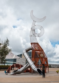 A modern playground featuring an elaborate climbing structure made of metal and geometric shapes dominates the scene. The structure includes multiple slides, with a futuristic design of intersecting loops and arcs. Surrounding the playground are trees and a paved area, with a large building in the background. A person stands near the playground, adding a sense of scale to the sculpture-like design.
