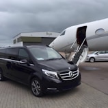 A black Mercedes-Benz van is parked on the tarmac next to a private jet with an open door and a staircase leading up. The setting is overcast, with a large hangar building labeled 'Harrods' in the background. Another white vehicle is partially visible to the right.
