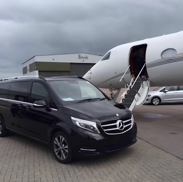 A black Mercedes-Benz van is parked on the tarmac next to a private jet with an open door and a staircase leading up. The setting is overcast, with a large hangar building labeled 'Harrods' in the background. Another white vehicle is partially visible to the right.