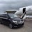 A black Mercedes-Benz van is parked on the tarmac next to a private jet with an open door and a staircase leading up. The setting is overcast, with a large hangar building labeled 'Harrods' in the background. Another white vehicle is partially visible to the right.