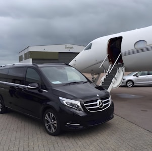 A black Mercedes-Benz van is parked on the tarmac next to a private jet with an open door and a staircase leading up. The setting is overcast, with a large hangar building labeled 'Harrods' in the background. Another white vehicle is partially visible to the right.