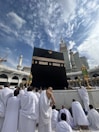 A group of people wearing white garments are gathered around the Kaaba, a large cuboid structure, against a backdrop of a partially cloudy sky. The Kaaba is adorned with intricate gold calligraphy and is set within a large mosque complex with several ornate structures and a clock tower in the background.