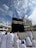 A group of people wearing white garments are gathered around the Kaaba, a large cuboid structure, against a backdrop of a partially cloudy sky. The Kaaba is adorned with intricate gold calligraphy and is set within a large mosque complex with several ornate structures and a clock tower in the background.