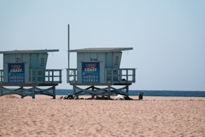 Lifeguard towers are positioned on a sandy beach with the ocean visible in the background. Each tower displays a blue sign promoting coastal care. The beach is flat and expansive, with a clear blue sky above.