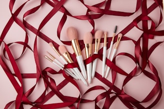 A collection of makeup brushes with white handles and rose gold accents is artistically arranged on a light pink surface. They are intertwined with elegant dark red satin ribbons, adding a touch of luxury and sophistication.