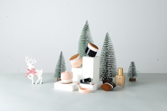 An arrangement of cosmetics is displayed on a light-colored surface, surrounded by small artificial pine trees dusted with white, evoking a wintery atmosphere. A decorative white reindeer with a red-checkered bow around its neck stands next to the setup, adding a festive touch.