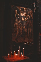 A religious icon depicting two haloed figures is displayed in a dimly lit setting. In the foreground, a collection of lit candles provides a soft, warm glow against the dark surroundings, enhancing the sacred and contemplative ambiance of the scene.