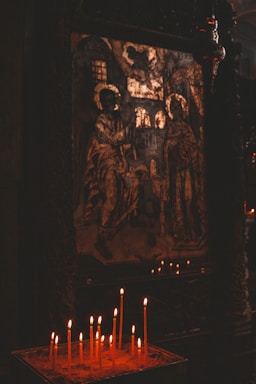A religious icon depicting two haloed figures is displayed in a dimly lit setting. In the foreground, a collection of lit candles provides a soft, warm glow against the dark surroundings, enhancing the sacred and contemplative ambiance of the scene.