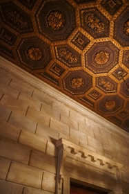 An interior view showcasing a richly decorated ceiling with intricate golden patterns set within hexagonal and rectangular frames. The walls are textured and constructed with large, evenly spaced stone-like bricks. A decorative door frame with ornate molding and detailing is visible near the bottom.