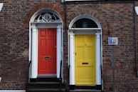 Two adjoining front doors, one painted red and the other yellow, are set against a brick facade with white columns and arched windows above each door. A sign indicating disabled parking is visible on the right side.