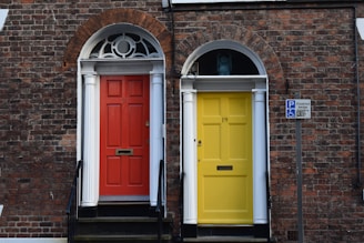 Two adjoining front doors, one painted red and the other yellow, are set against a brick facade with white columns and arched windows above each door. A sign indicating disabled parking is visible on the right side.