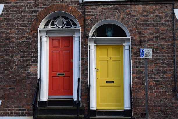 Two adjoining front doors, one painted red and the other yellow, are set against a brick facade with white columns and arched windows above each door. A sign indicating disabled parking is visible on the right side.