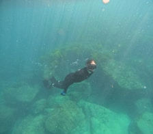 A person wearing snorkeling gear swims underwater near large rocks. The blue-green water creates a clear view of the surrounding environment, with sunlight streaming through the surface and illuminating the scene.