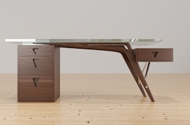 A modern wooden desk with a unique, artistic design features three drawers on the left side and one on the right. The structure includes elegant curves and a glass top. The desk is set against a simple, light-colored background on a wooden floor.