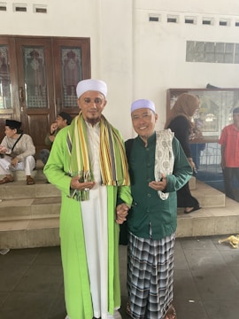 Two men are standing together in traditional attire, smiling and holding hands. The man on the left is wearing a bright green robe with a white headpiece and a multicolored scarf, while the man on the right is in a darker green outfit with a light lavender cap. Other people are visible in the background, sitting on tiled steps, indicating a social or religious gathering.