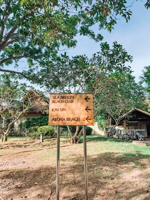 A signpost with directional arrows is situated outdoors in a lush, green area. The sign provides directions to 'Sea Breeze Beach Club,' 'Kai Spa,' and 'Alona Beach.' The signpost is positioned in a landscaped area with trees, a house or building in the background, and some bicycles parked to the right.