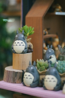 Several Totoro figurines are arranged on wooden platforms surrounded by small succulent plants. The scene appears to be part of a display setup, possibly in a store or a themed exhibition, with soft and muted lighting adding a cozy atmosphere.