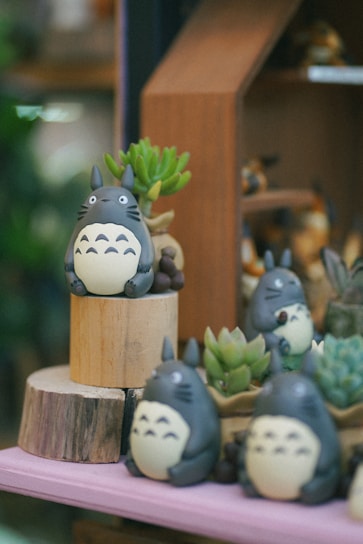 Several Totoro figurines are arranged on wooden platforms surrounded by small succulent plants. The scene appears to be part of a display setup, possibly in a store or a themed exhibition, with soft and muted lighting adding a cozy atmosphere.
